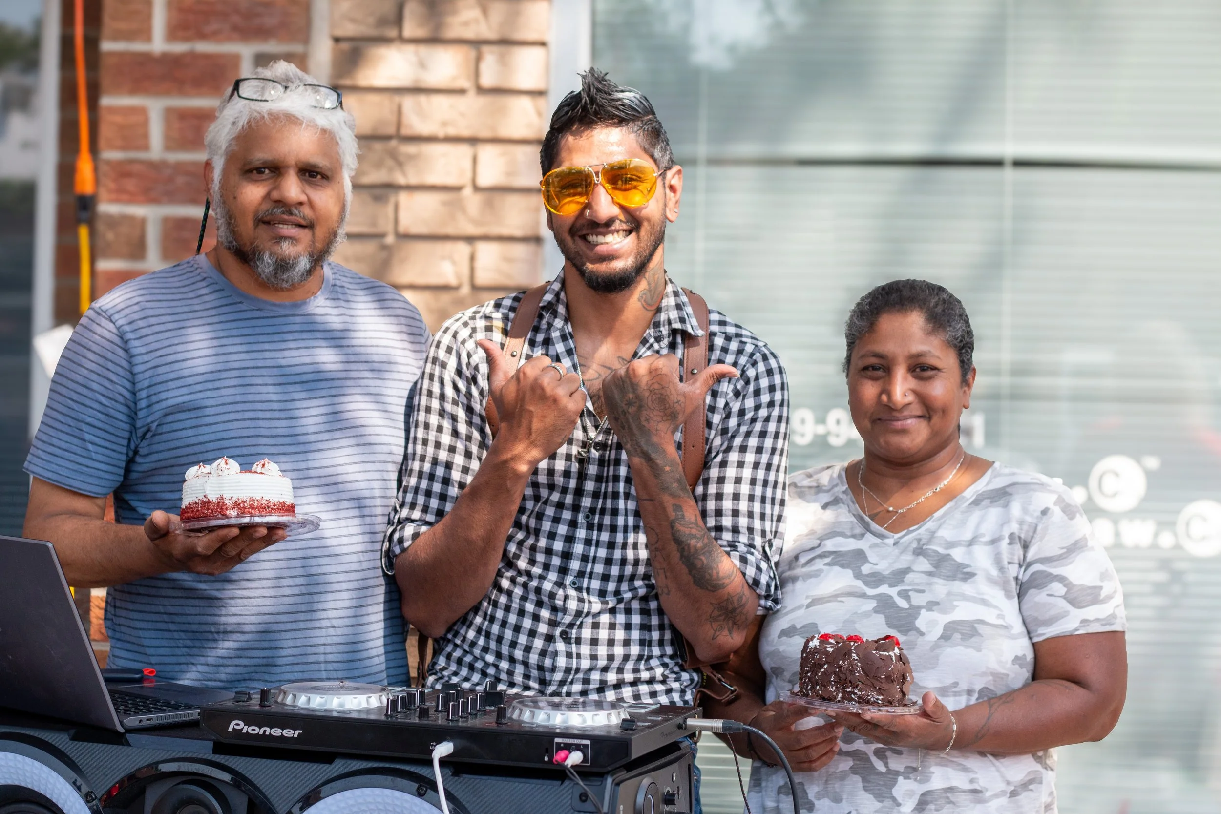 Three people standing outdoors, smiling, holding cakes, with DJ equipment in front of them. One man has gray hair and a beard, another man has dark hair, sunglasses, and tattoos, and a woman has short curly hair; all appear to be celebrating a birthd