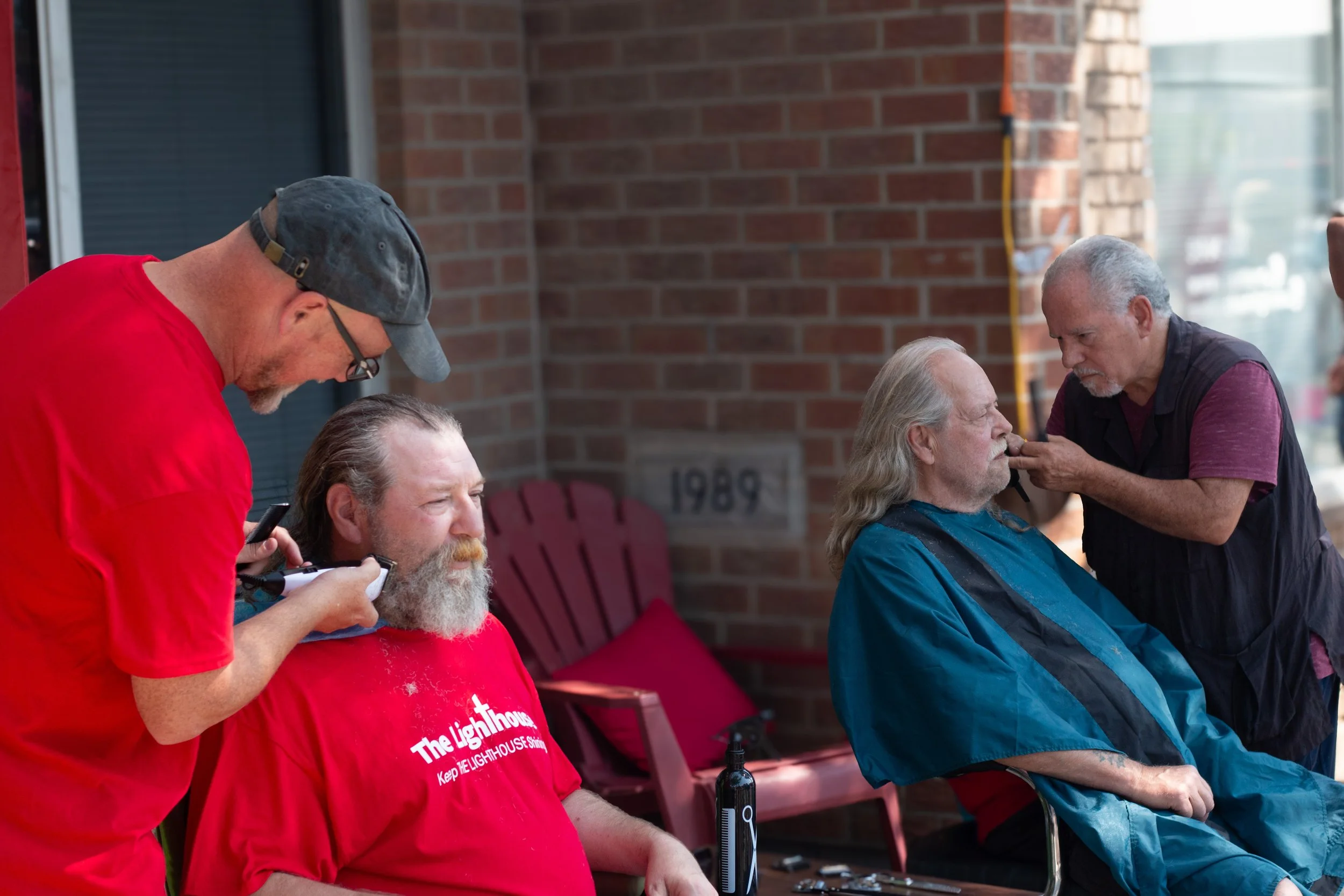 Two men getting haircuts outdoors near a brick building. One man has long gray hair and a beard, sitting in a chair wearing a blue cape. The barber is working on his beard. A second man with gray hair and a beard in a red t-shirt sits nearby. The barber is focused on trimming the first man's beard.