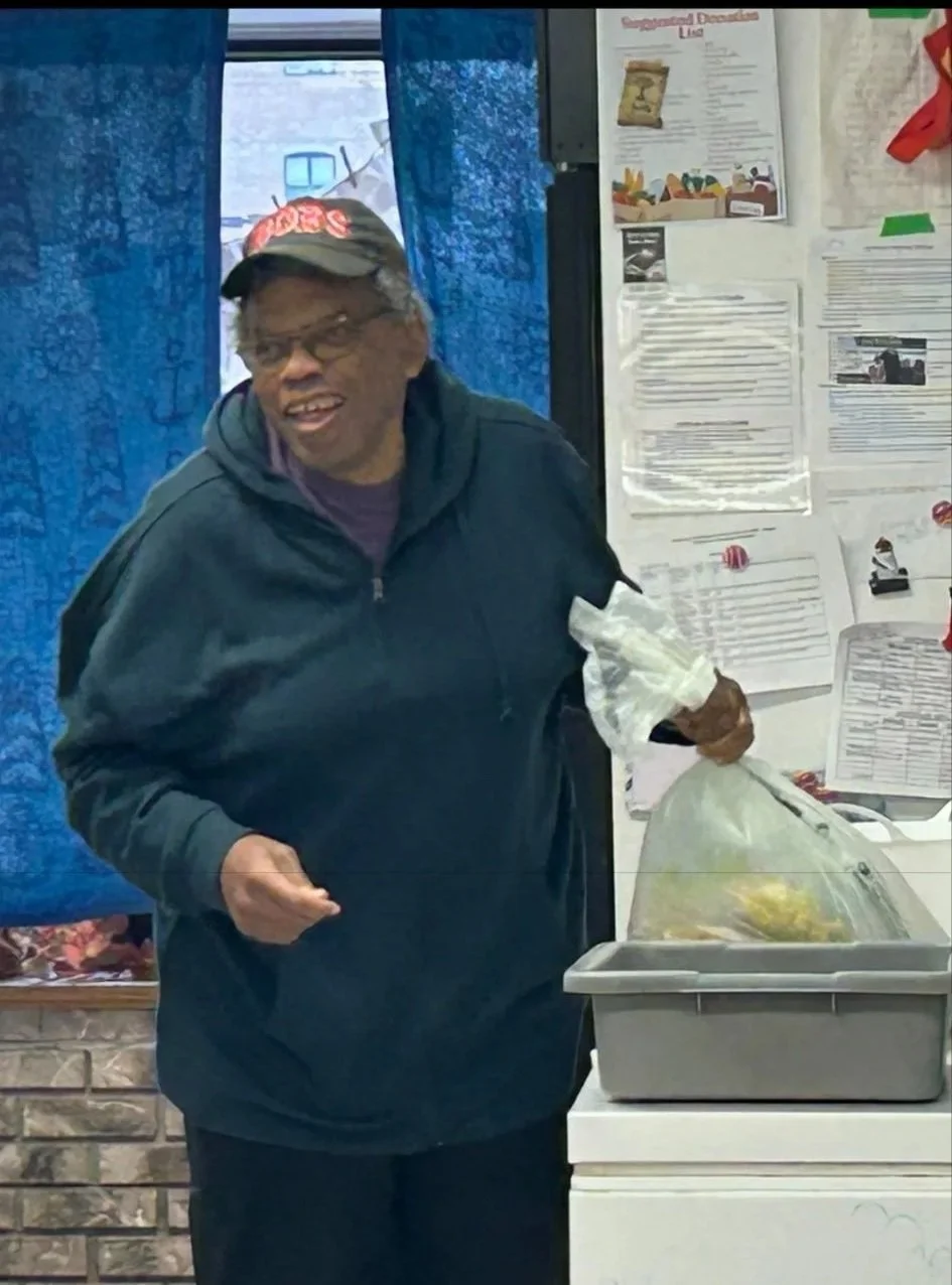A woman smiling, wearing a black hoodie and a black cap, standing at a food service or donation station with a tray and a plastic bag.