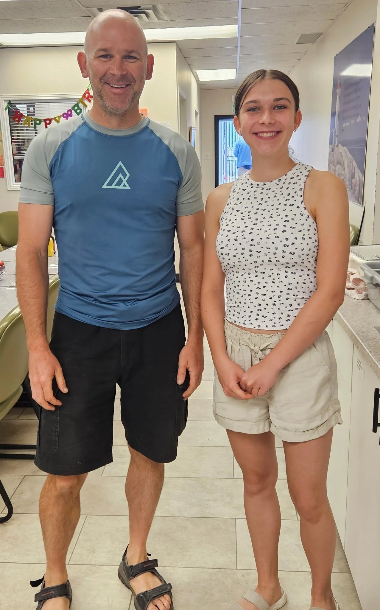 A man and a woman stand side by side indoors, smiling at the camera. The man is wearing a blue and gray athletic shirt, black shorts, and sandals. The woman is wearing a sleeveless white top with black patterns, beige shorts, and sandals. The background features a banner with