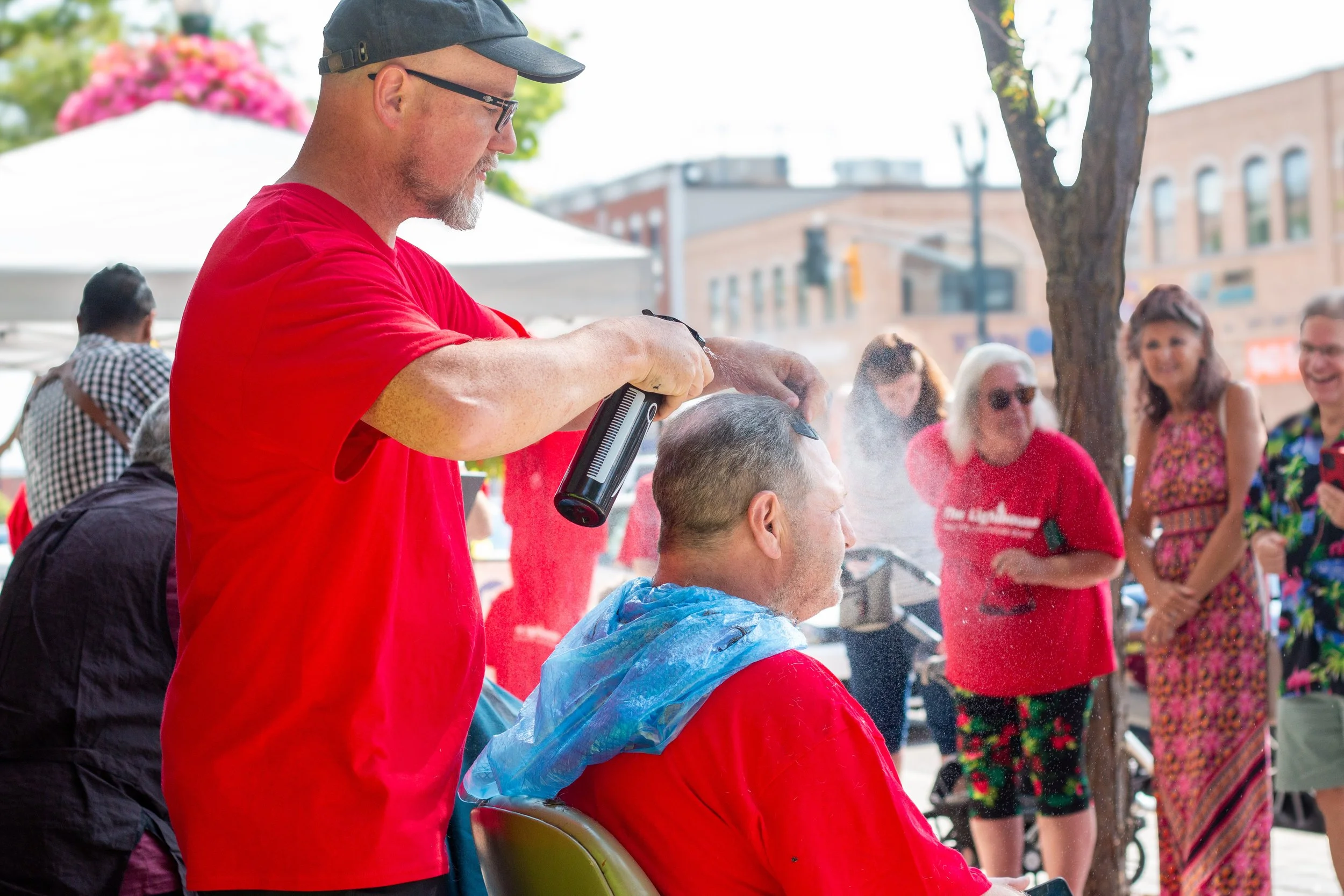 Man in red T-shirt getting a haircut outdoors while people watch and laugh, some wearing red shirts, on a city street with trees and buildings in the background.