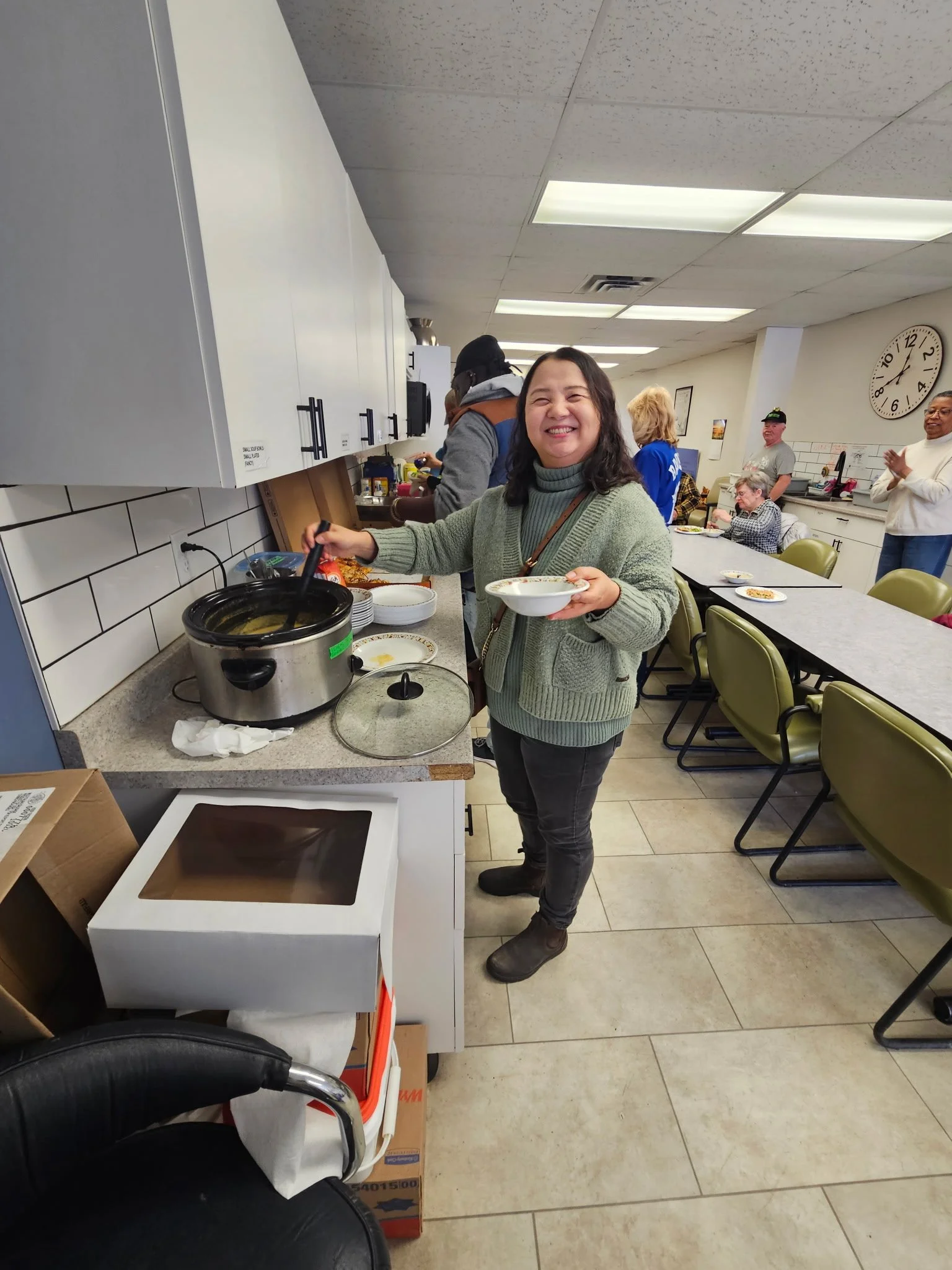 A woman smiling and serving food in a soup kitchen or community dining area, with other people in the background doing the same.