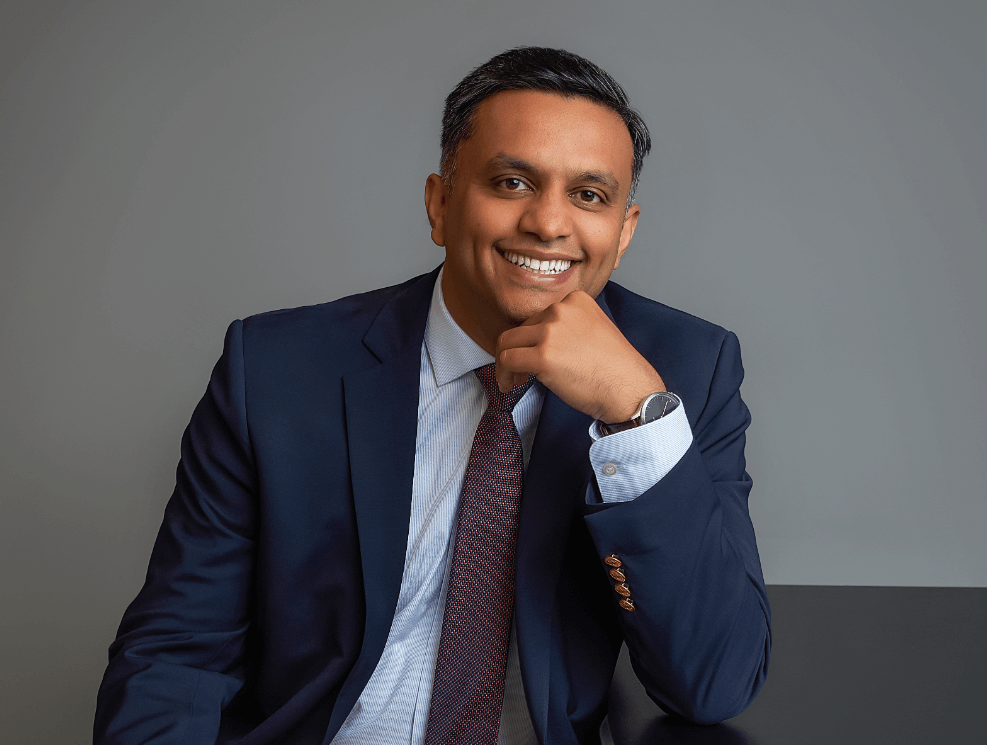 Professional man in navy suit with light blue shirt and dark red tie smiling and resting his chin on his hand against a gray background.