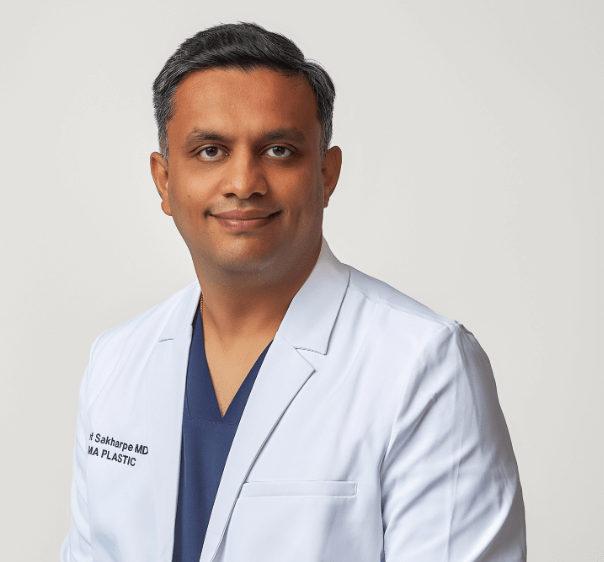 A male doctor wearing a white coat and blue scrubs, smiling at the camera against a plain light background.