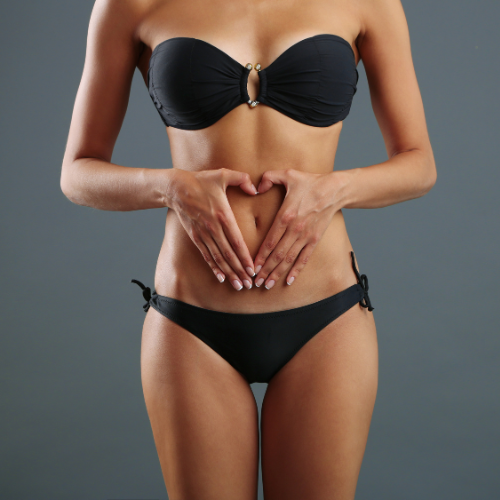 A woman wearing a black bikini, standing against a gray background, with her hands forming a heart shape over her stomach.