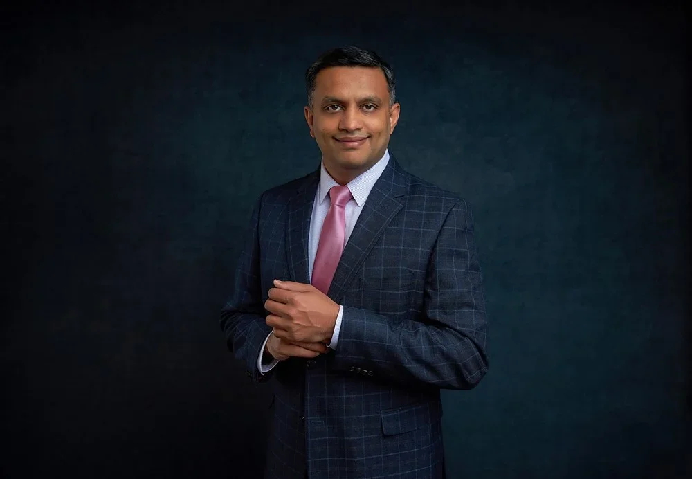 Portrait of a man in a dark checkered suit with a pink tie, standing against a dark background.