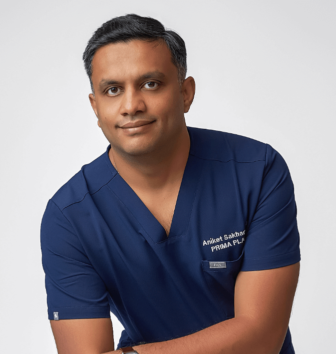 Portrait of a man wearing blue medical scrubs with the name Aniket Sakhar and PRIMA PLASTIC SURGERY embroidered on it, against a plain white background.