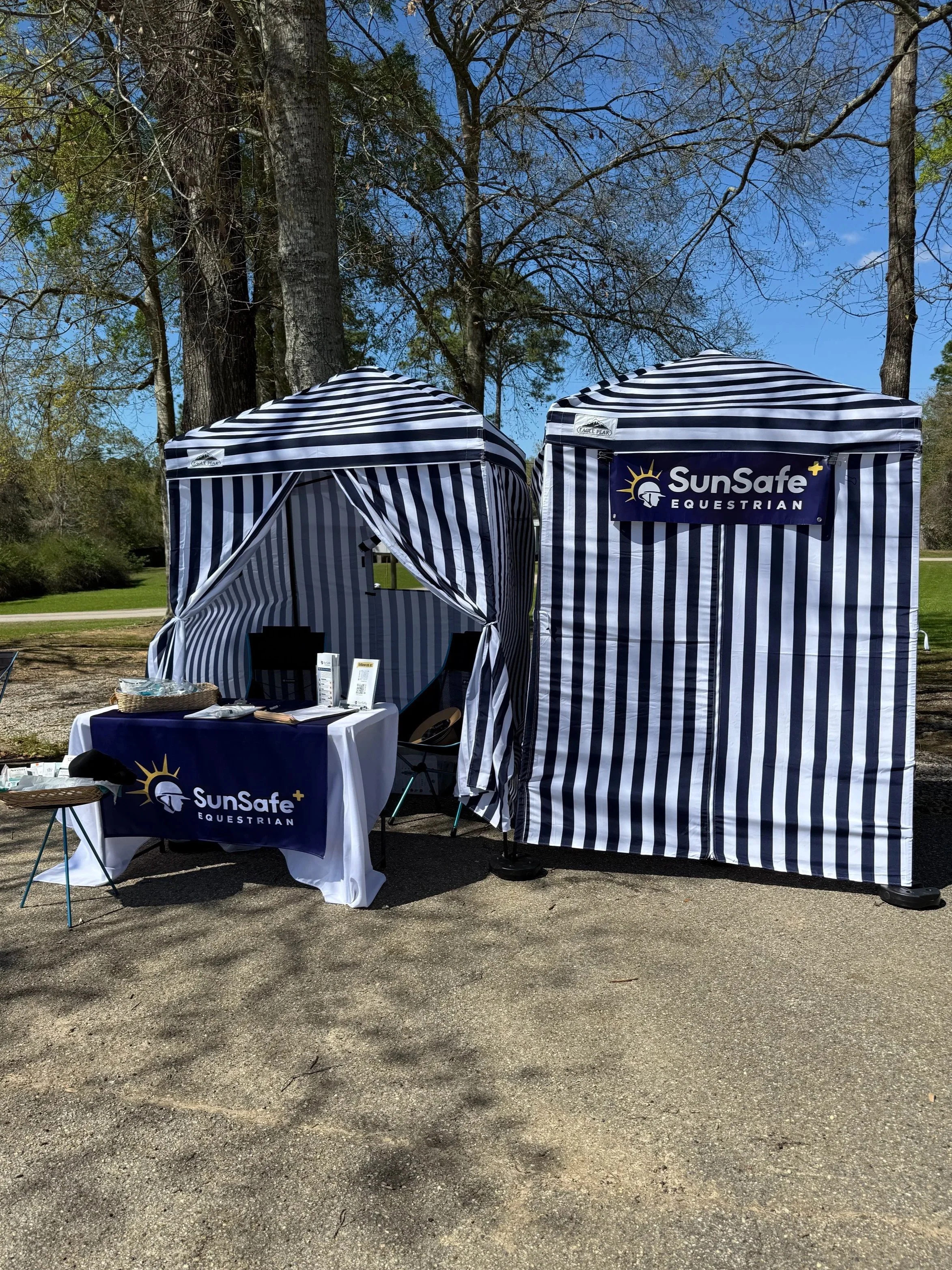 Outdoor event tents and a table with promotional material for SunSafe Equestrian, set up on a paved area with trees in the background.