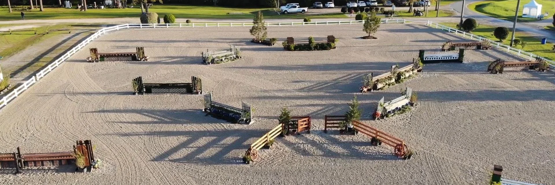 An outdoor equestrian riding arena with various jumps and obstacles set up, surrounded by a white fence and some trees.