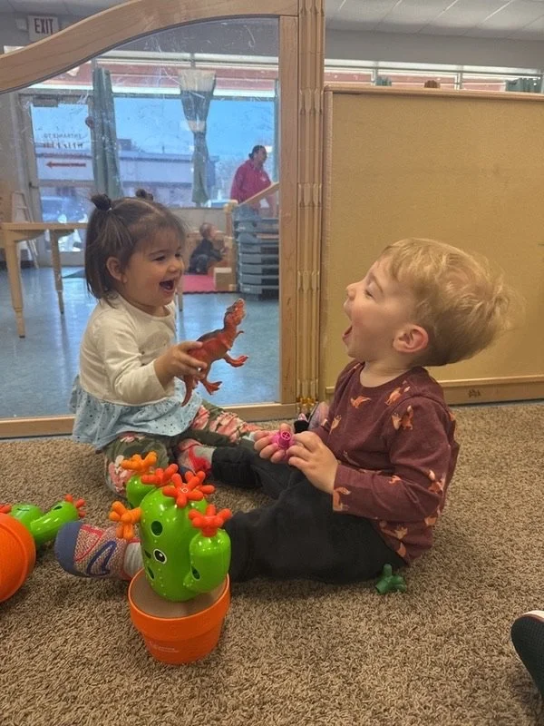Two young children, a girl and a boy, sitting on the carpet playfully pretending to have a lively interaction, with the girl holding a toy lobster and both children laughing.