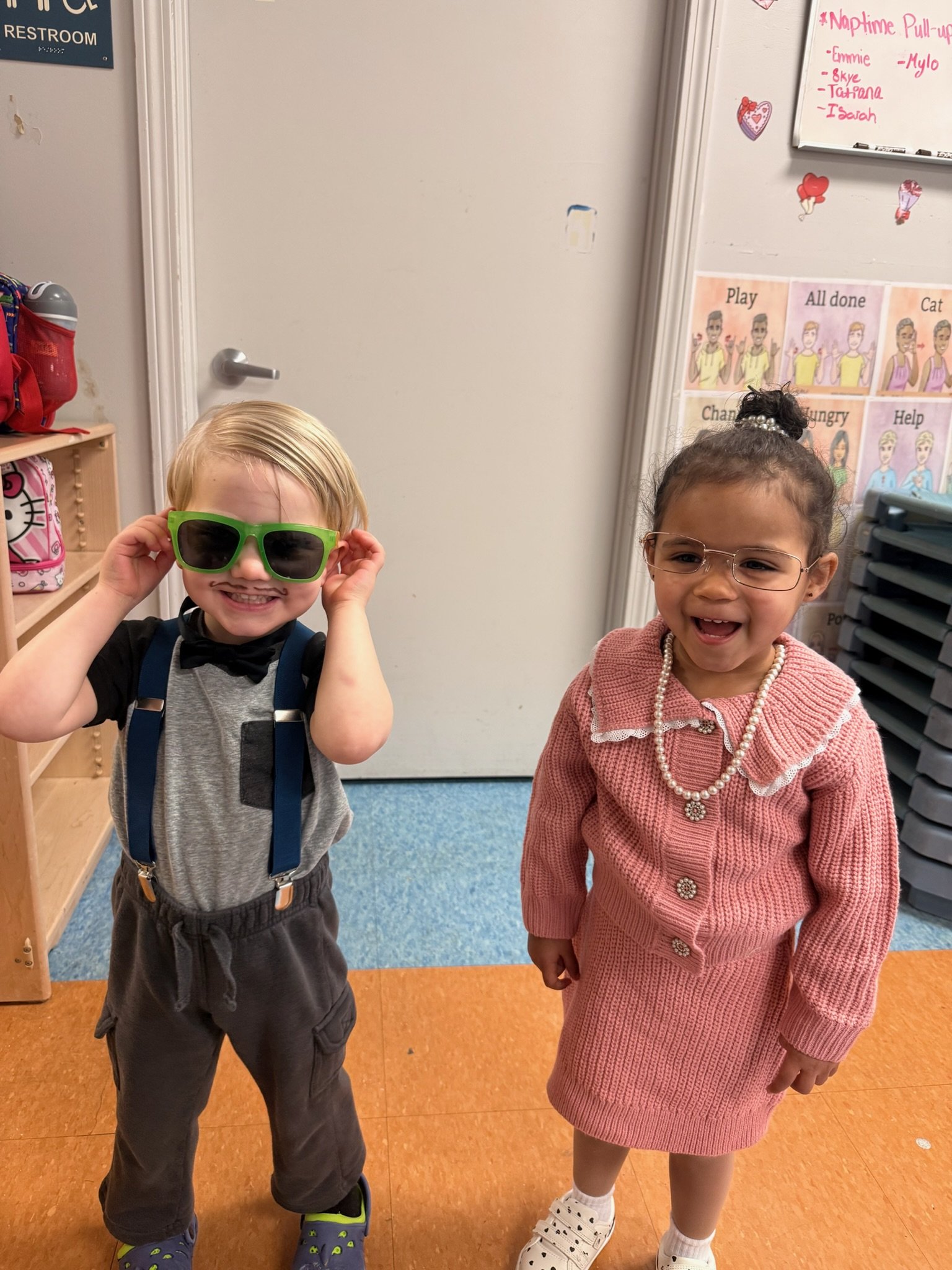 Two young children standing in a room, smiling at the camera. The boy on the left is wearing green sunglasses, a gray shirt with suspenders, dark pants, and colorful shoes. The girl on the right is wearing glasses, a pink cardigan, a pearl necklace, and white shoes. The background includes a white door, a poster with illustrations, and some wall decorations.