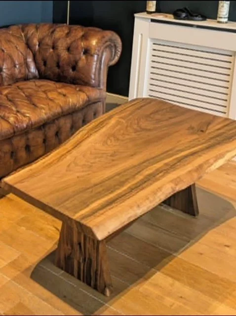 Wooden coffee table with a natural finish in a living room setting. A brown leather tufted sofa and a white radiator cover are visible in the background.