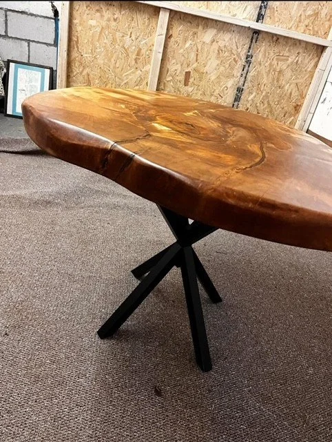 A wooden table with a thick, polished, live edge surface and black metal cross-legged base in a workshop setting.