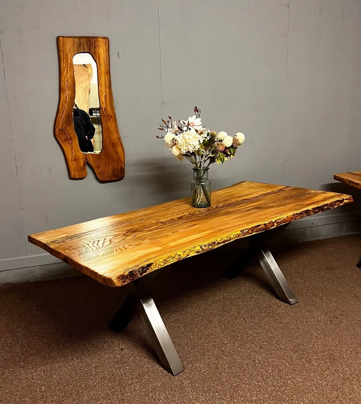 A rustic wooden table with metal legs, decorated with a glass vase of white and pink flowers, in a room with gray walls and a small wooden mirror hanging on the wall.