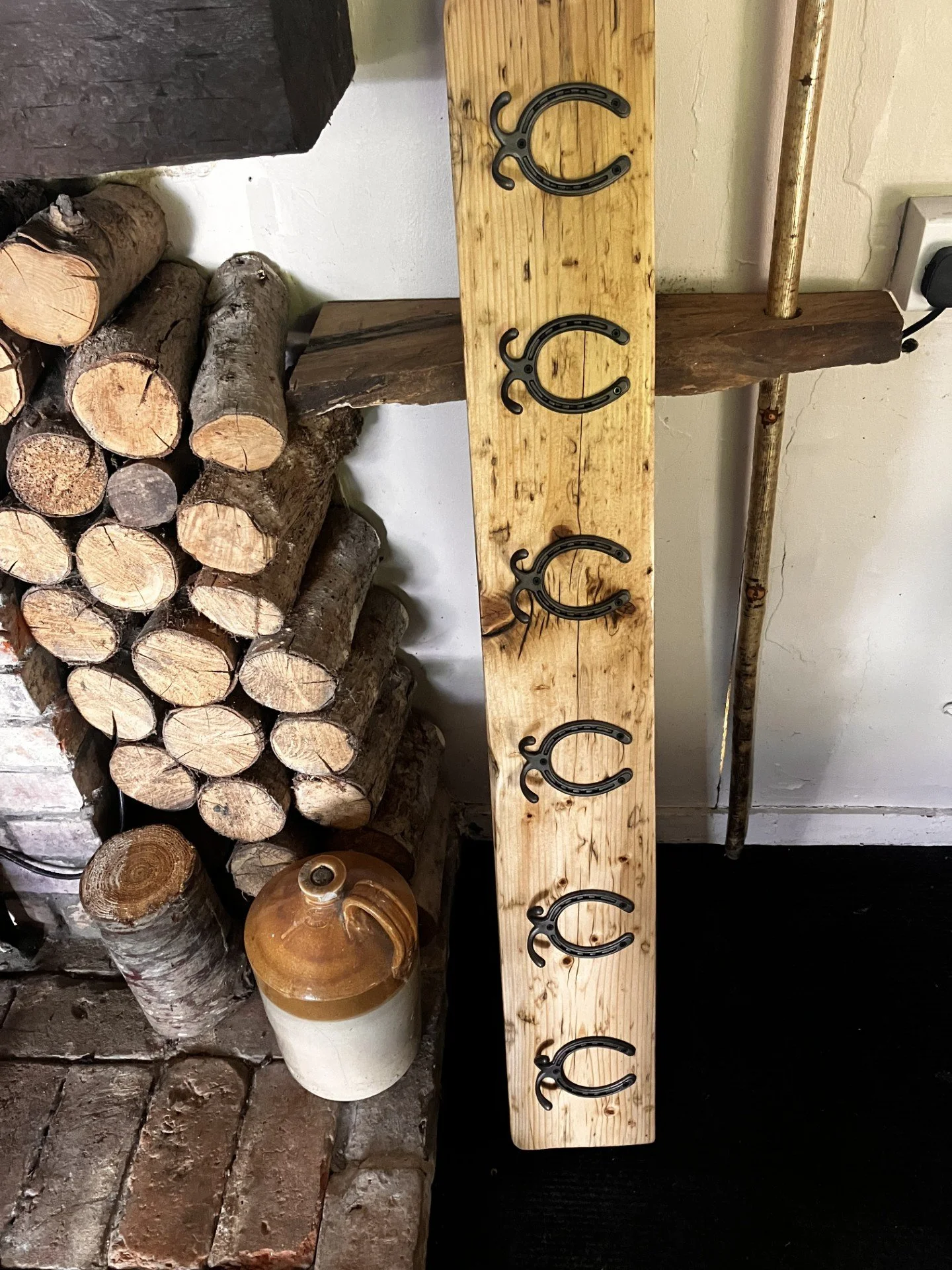A wooden board with five horseshoes attached vertically, surrounded by stacked logs on one side and a ceramic jug on a brick surface, set against a wall with a power outlet.