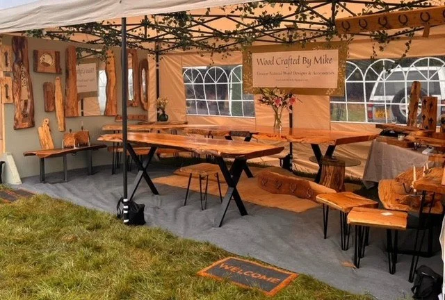 Woodcrafted furniture display inside a tent with wooden tables and benches, wall art, and a sign reading 'Wood Crafted By Mike' at an outdoor market.