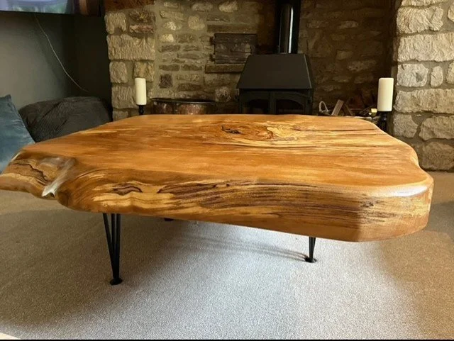 A wooden coffee table with a smooth, polished surface and irregular shape, supported by black metal legs, situated on a carpeted floor in a room with stone walls and a black stove.