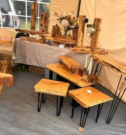 Display of rustic wooden furniture, including tables and shelves, with a floral arrangement on a table in a tent-like setting.