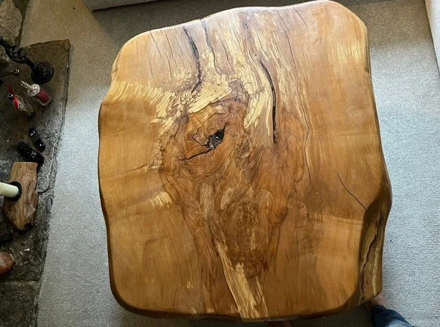 A square wooden table with a natural, uneven edge, showing a prominent knot and grain pattern in the center.