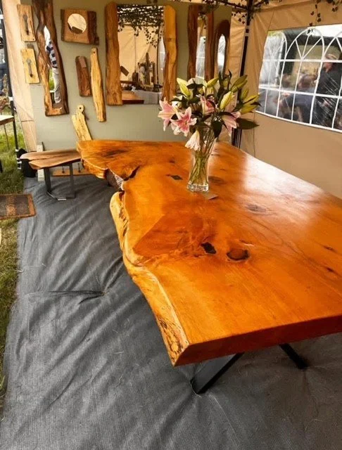 A large, natural wood slab table with a glossy finish, decorated with a bouquet of pink and white lilies in a glass vase, inside a tent with wooden decor on the walls and a window showing outdoor scenery.