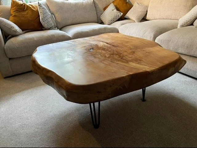 A wooden coffee table with a natural, uneven edge and metal hairpin legs in a living room with a beige sectional sofa and decorative pillows.