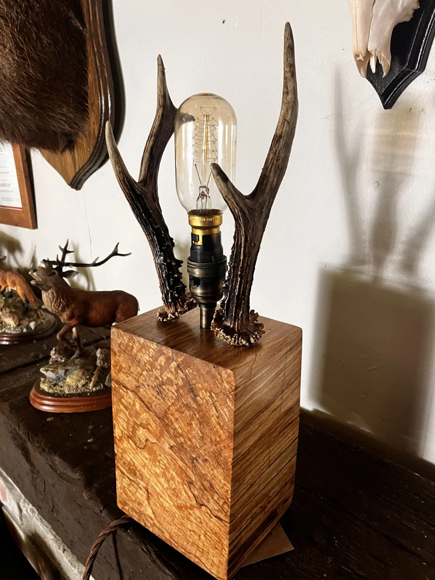 A decorative table lamp with an exposed bulb, mounted on a block of wood, supported by antlers, in a rustic style.