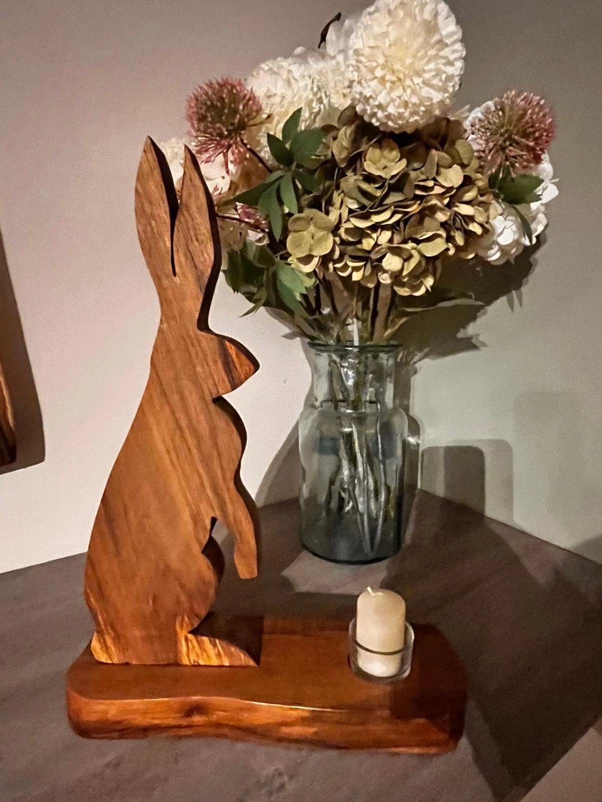 Decorative wooden hare sculpture, glass vase with dried white, pink, and green flowers, and a small candle on a wooden surface.