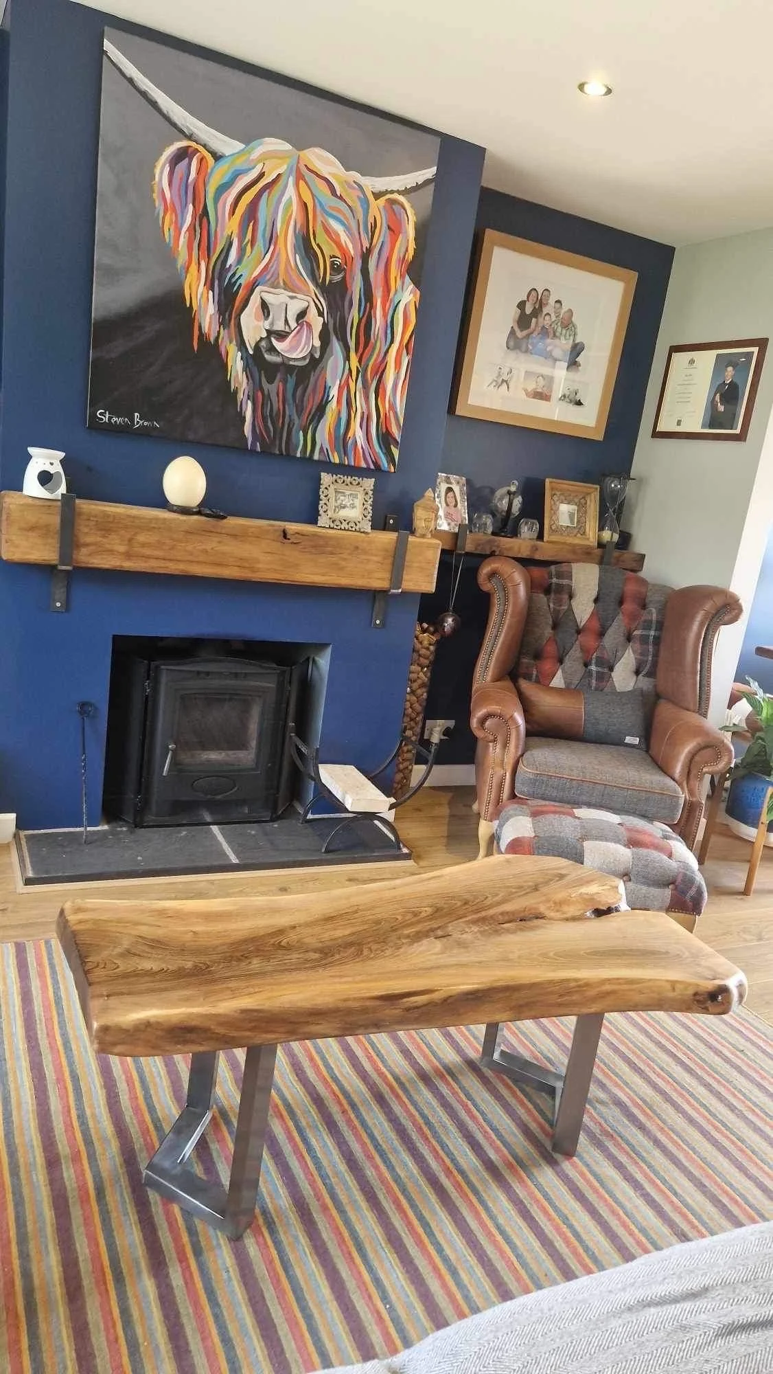 A cozy living room with a colorful abstract painting of a Highland cow hanging above a wood-burning stove. There is a wooden mantel with framed photos and decorative items. To the right, an upholstered armchair with a matching ottoman features a patc