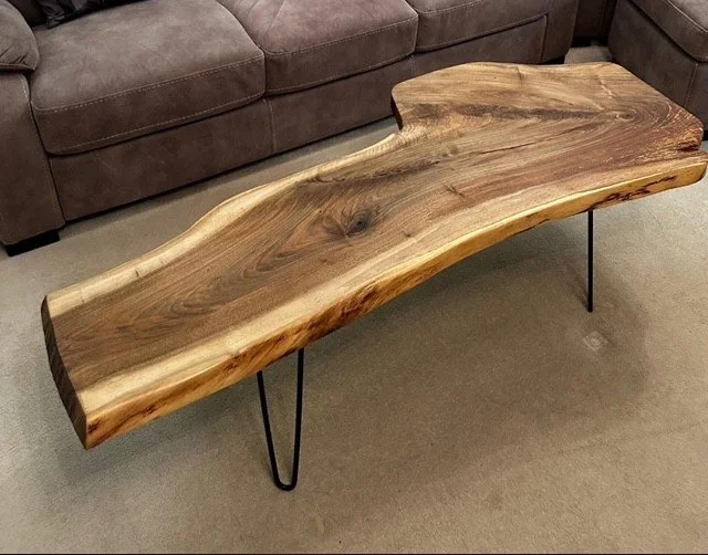 A wooden coffee table with a natural, irregular shape and visible wood grain, supported by black metal hairpin legs, placed in front of a brown sofa.