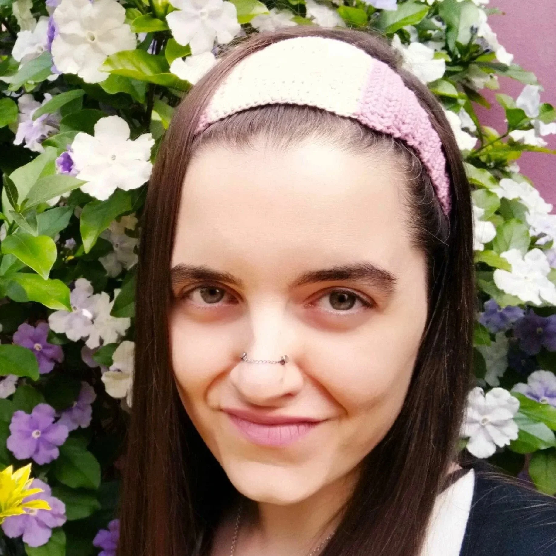 A woman with fair skin and long brown hair, wearing a pink and white headband, smiling, standing in front of a background of white, purple, and yellow flowers.