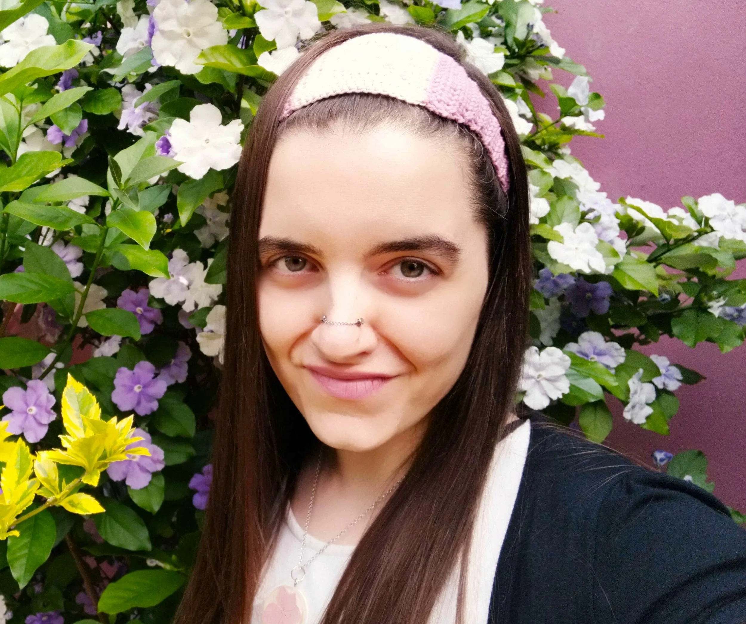 A young woman with long brown hair, wearing a pink and purple headband and a black and white top, standing in front of a bush with white, purple, and yellow flowers.