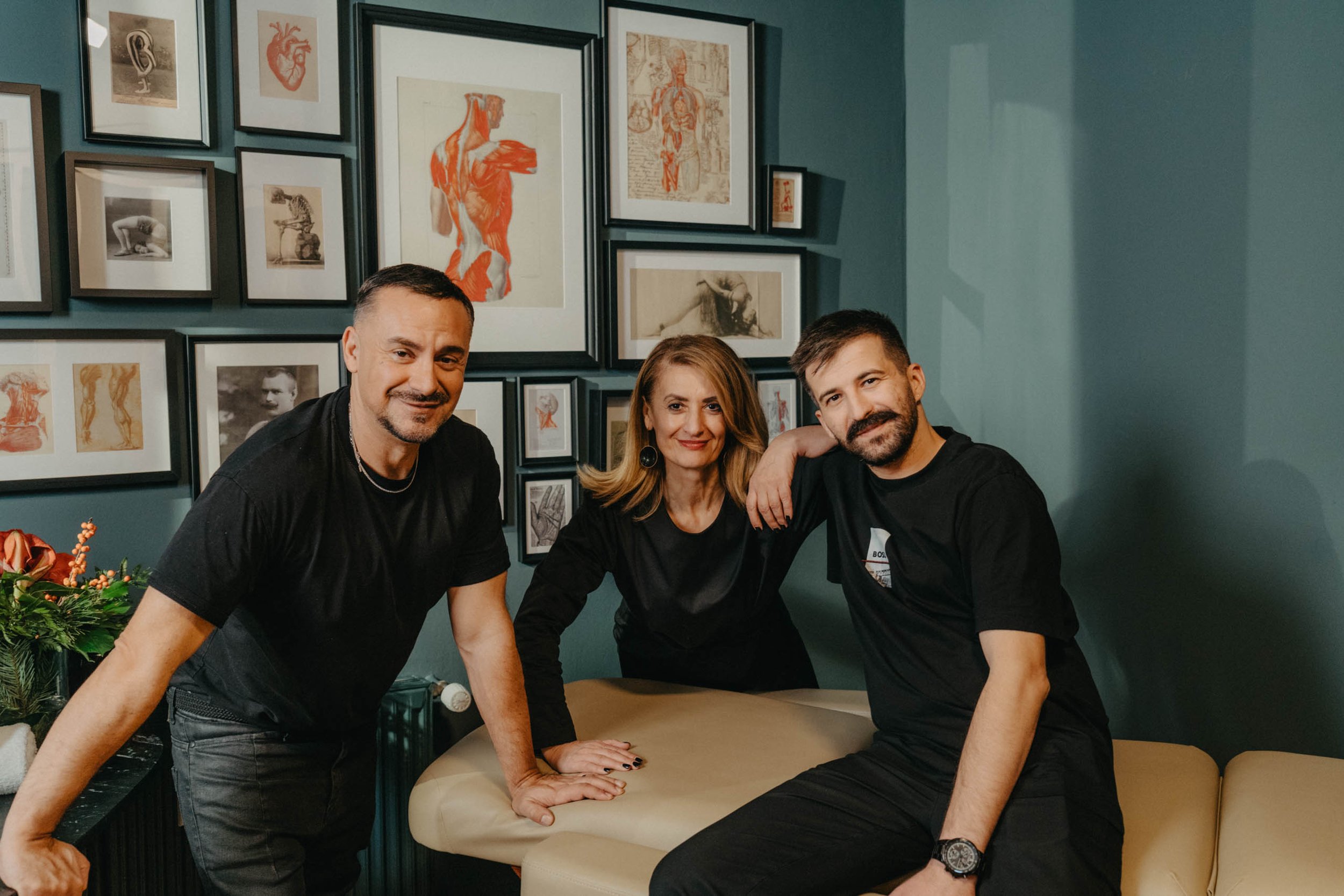 Drei Personen, zwei Männer und eine Frau, posieren in einem Wohnzimmer vor einer Wand mit mehreren Kunstbildern, die auf dunkelgrüner Wand hängen.