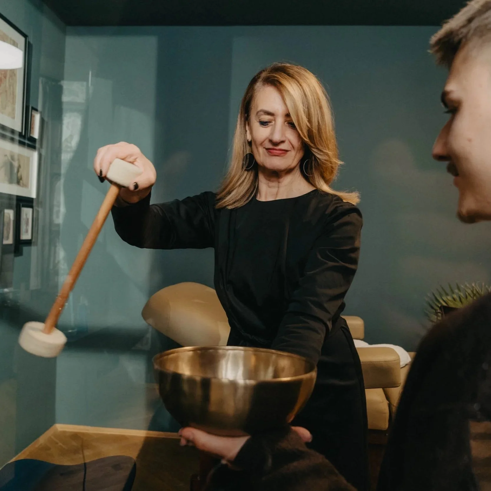 Frau schlägt mit einem Hammer auf eine Klangschale, während ein junger Mann die Schale hält.