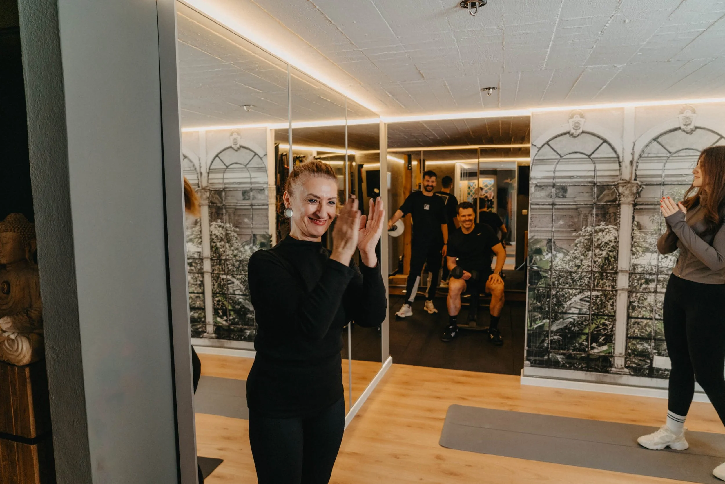 Ältere Frau applaudiert in einem Fitnessstudio, während junge Sportler im Hintergrund lachen und sich entspannen.