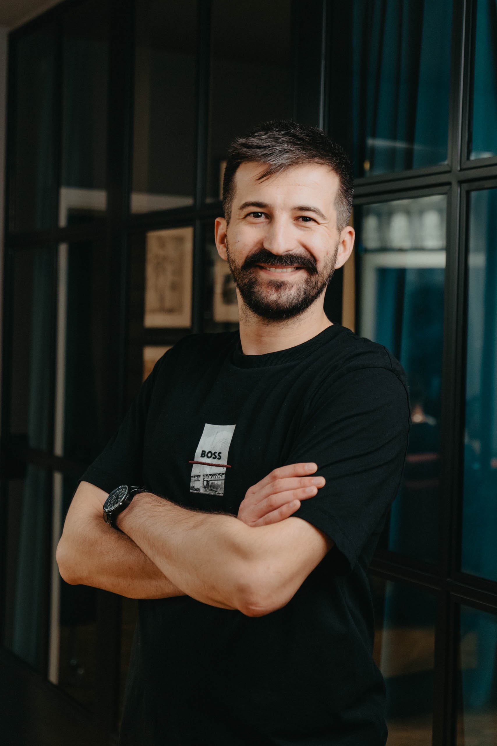 Ein lächelnder Mann mit dunklem Bart und kurzen Haaren, trägt ein schwarzes T-Shirt mit einem weißen Rechteck-Design sowie einem schwarzen Armband und steht vor einer schwarzen Wand mit Glasfenstern.