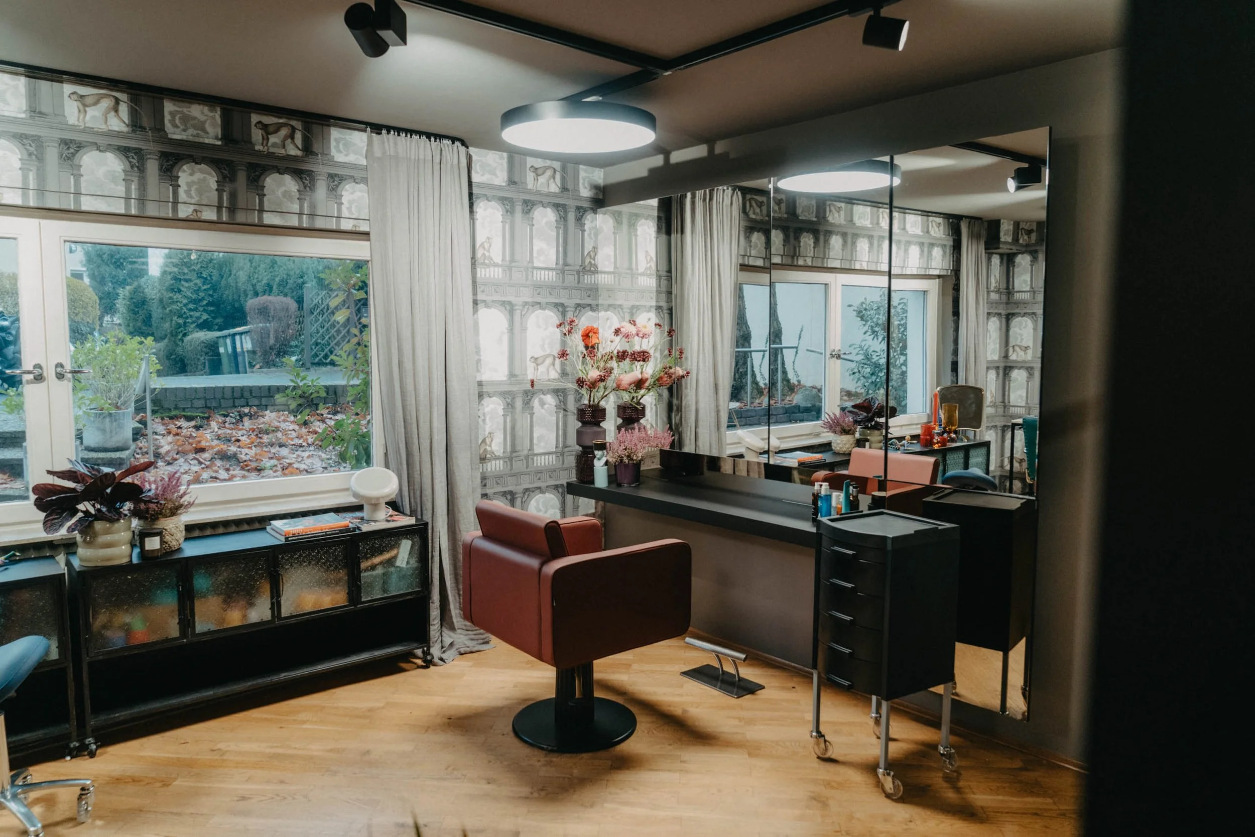Innenansicht eines stilvollen Friseursalons mit großen Fenstern, einem Spiegel, Friseurstuhl und Dekorationen mit Blumen.