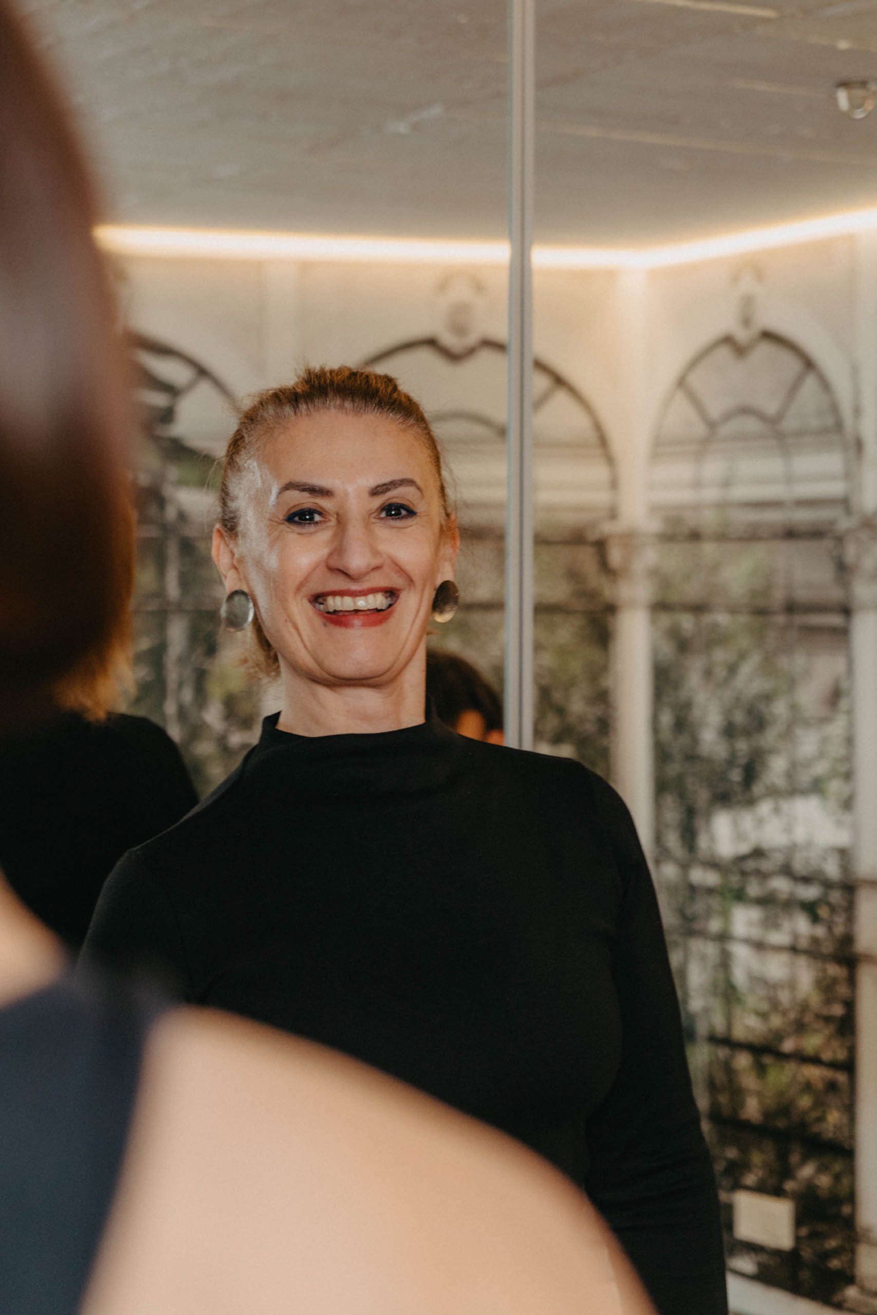 Eine lächelnde Frau in einem schwarzen Oberteil vor einem Spiegel, im Hintergrund ein festlich dekorierter Raum.