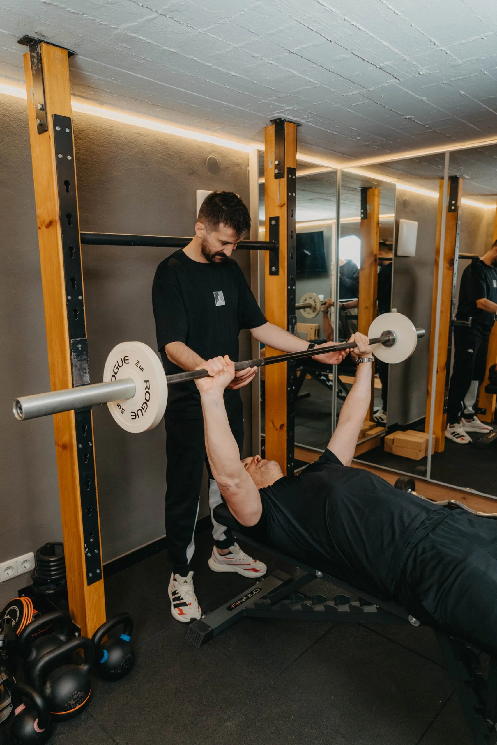 Eine Person liegt auf einer Bank und hebt eine Langhantel beim Krafttraining, während eine Aufpassperson daneben steht und beobachtet.