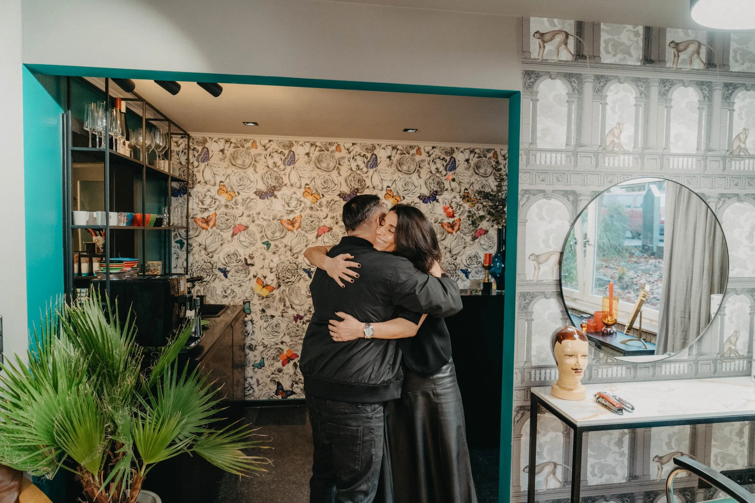 Ein Mann und eine Frau umarmen sich in einem gemütlichen Raum, umgeben von Pflanzen und kunstvollen Dekorationen.