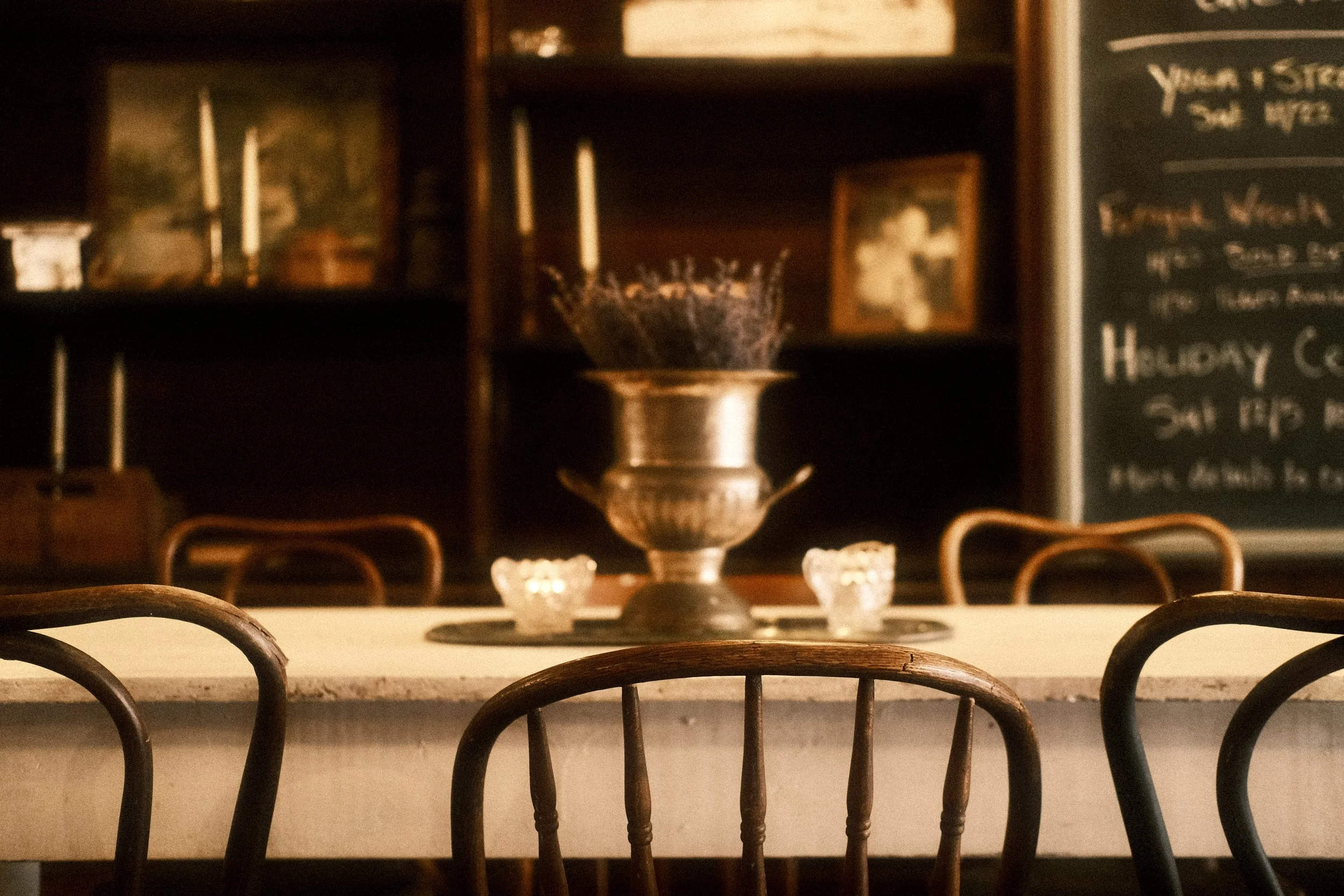 Empty dining table with chairs, centered decorative urn with purple flowers, and small candle holders, in a cozy, dimly lit restaurant or cafe with wooden shelves and a chalkboard menu in the background.