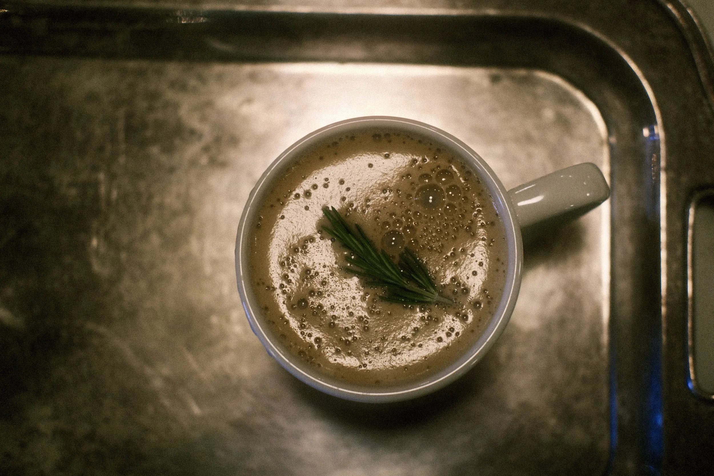 Top-down view of a white mug filled with frothy coffee, garnished with a sprig of rosemary, sitting on a metal sink countertop.