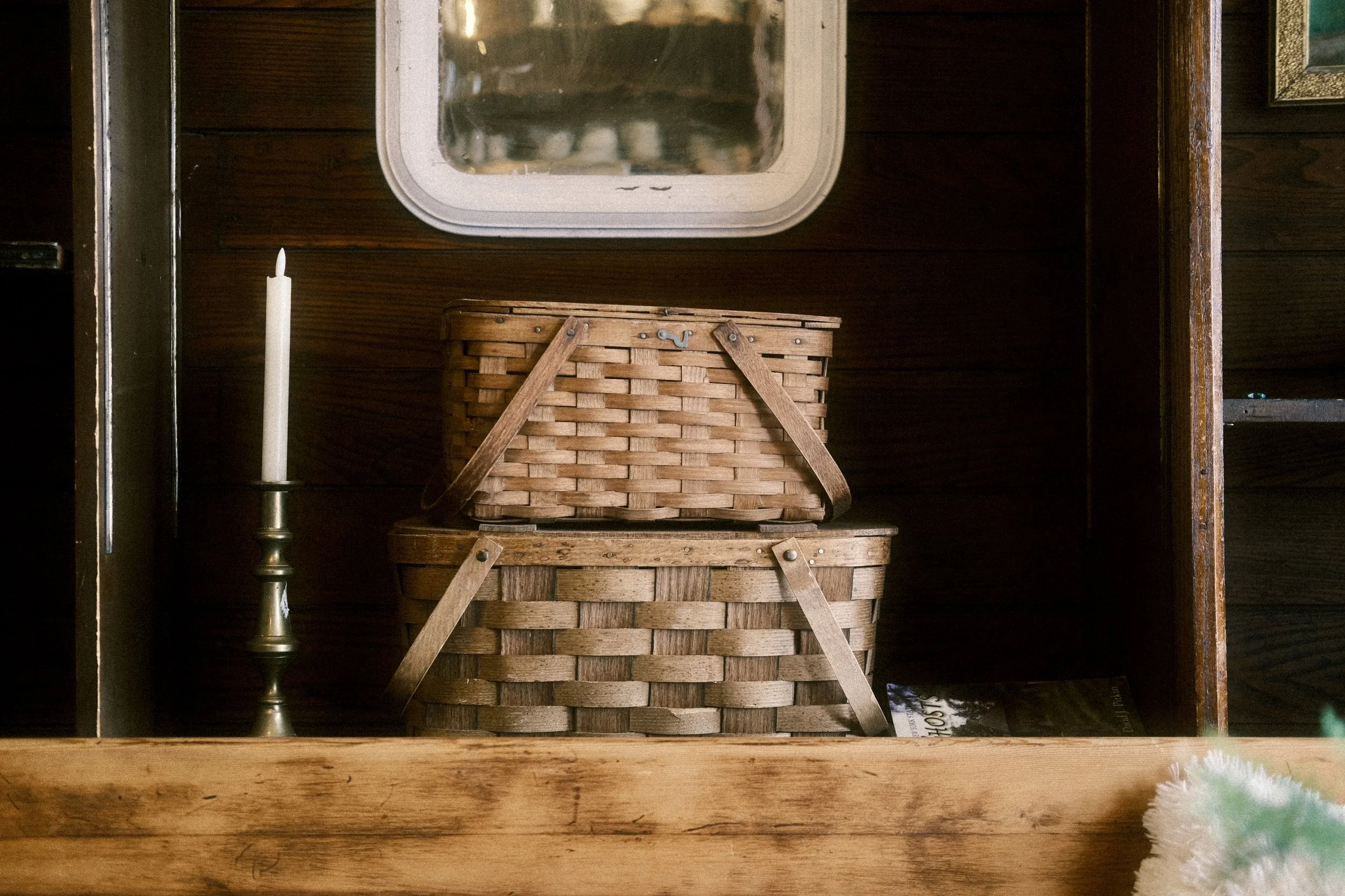 Two woven wooden baskets stacked inside a wooden cabinet, with a brass candlestick holding a white candle to the left and a mirror above.