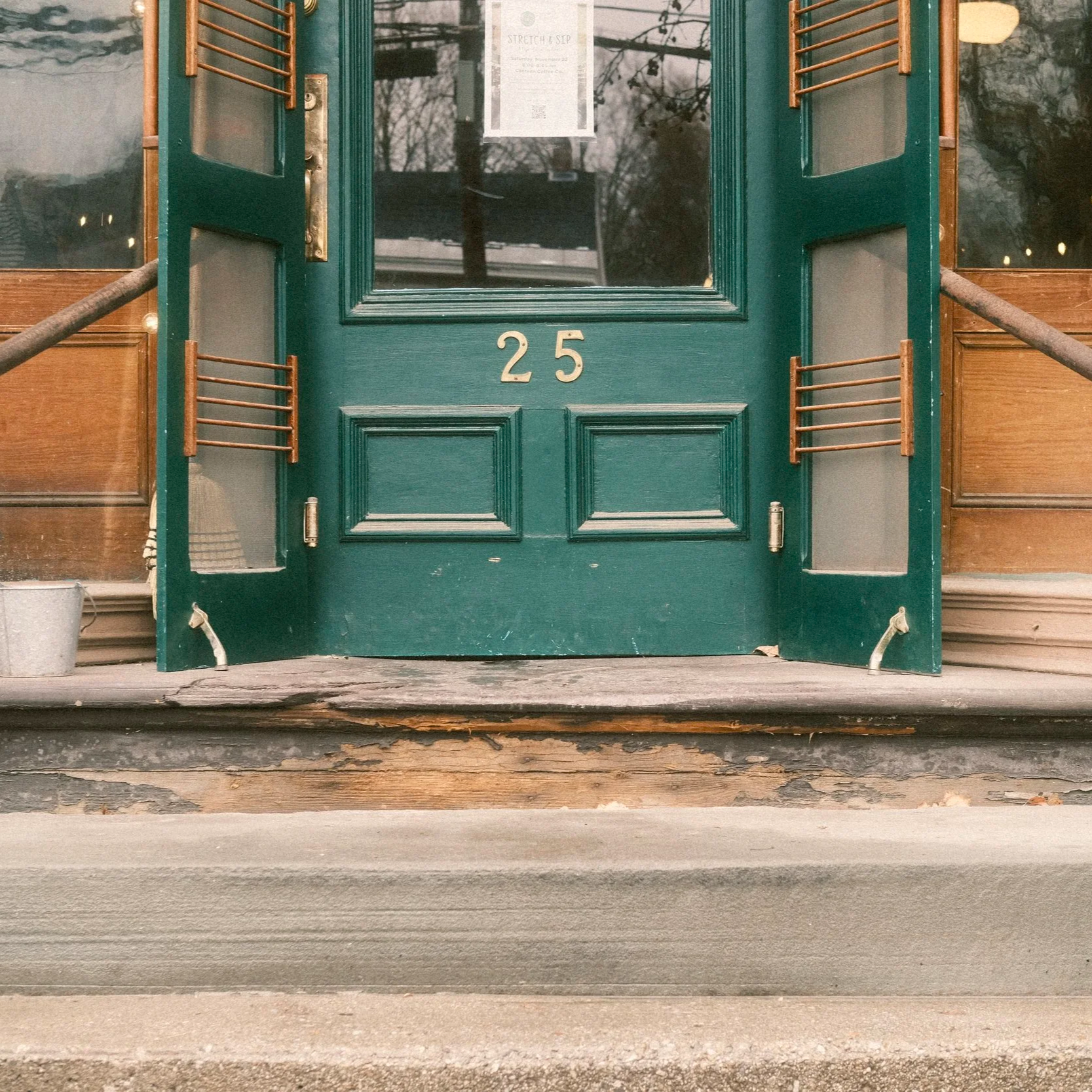 Close-up of a green front door with the number 25, open wooden shutters, a notice on the glass, and weathered wooden steps leading up to it.