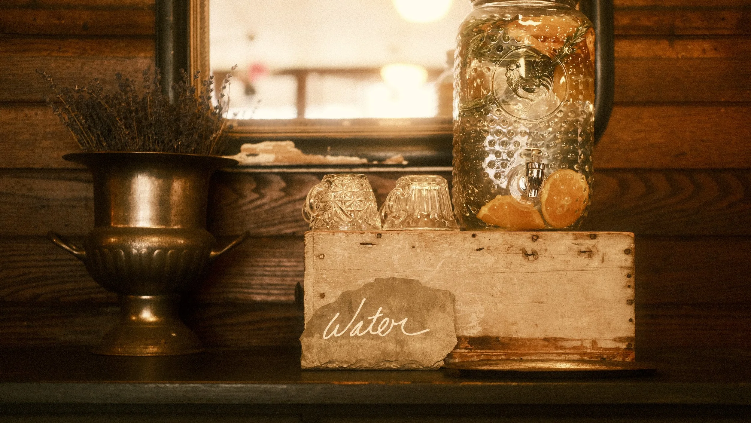 A rustic bar setup with a glass water dispenser containing lemon slices and mint leaves, two glass cups, a wooden box labeled 'Water', a brass vase with dried lavender, and a mirror reflected in the background
