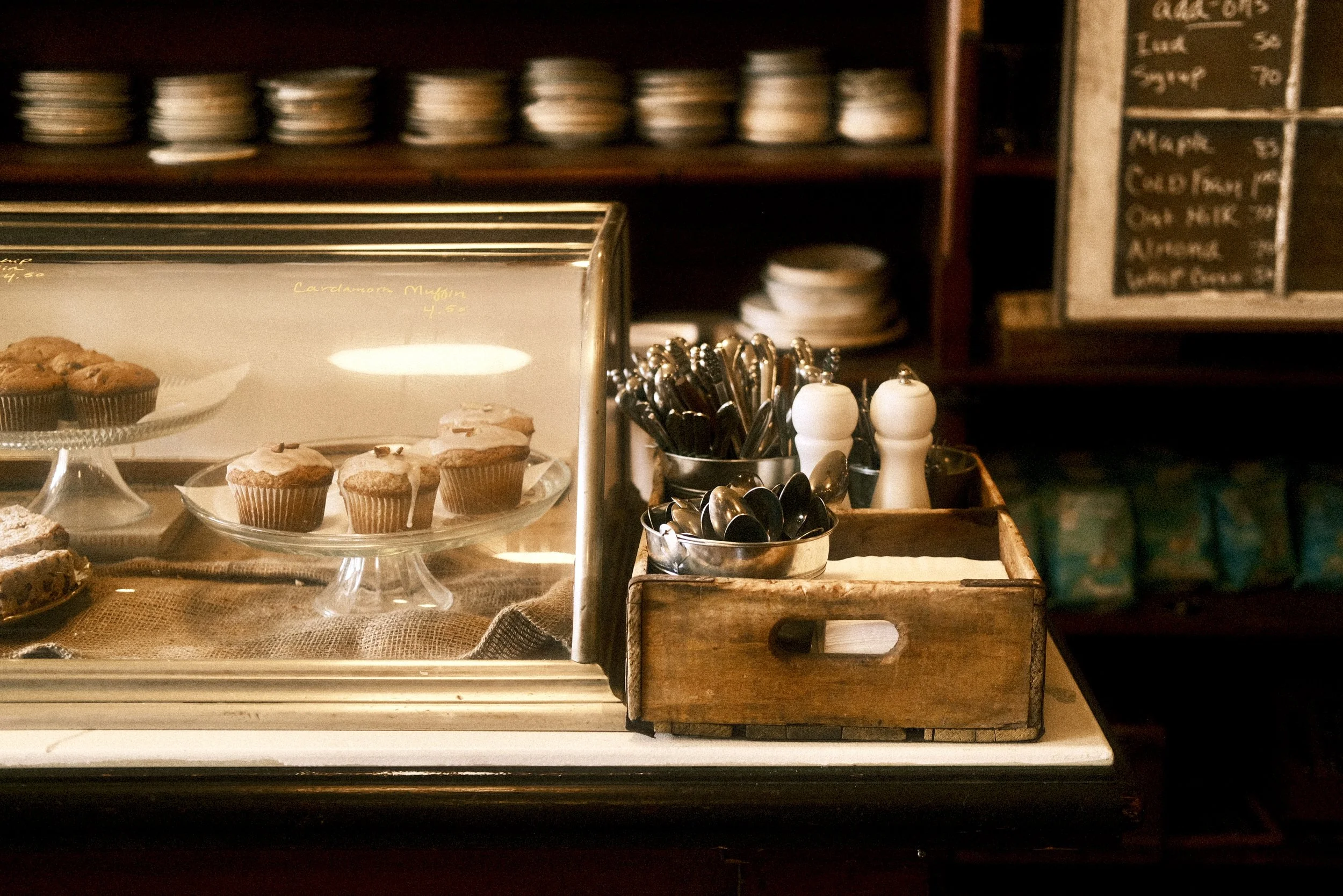 Display case with muffins and cupcakes, utensils, and condiments in a cozy cafe or bakery setting.