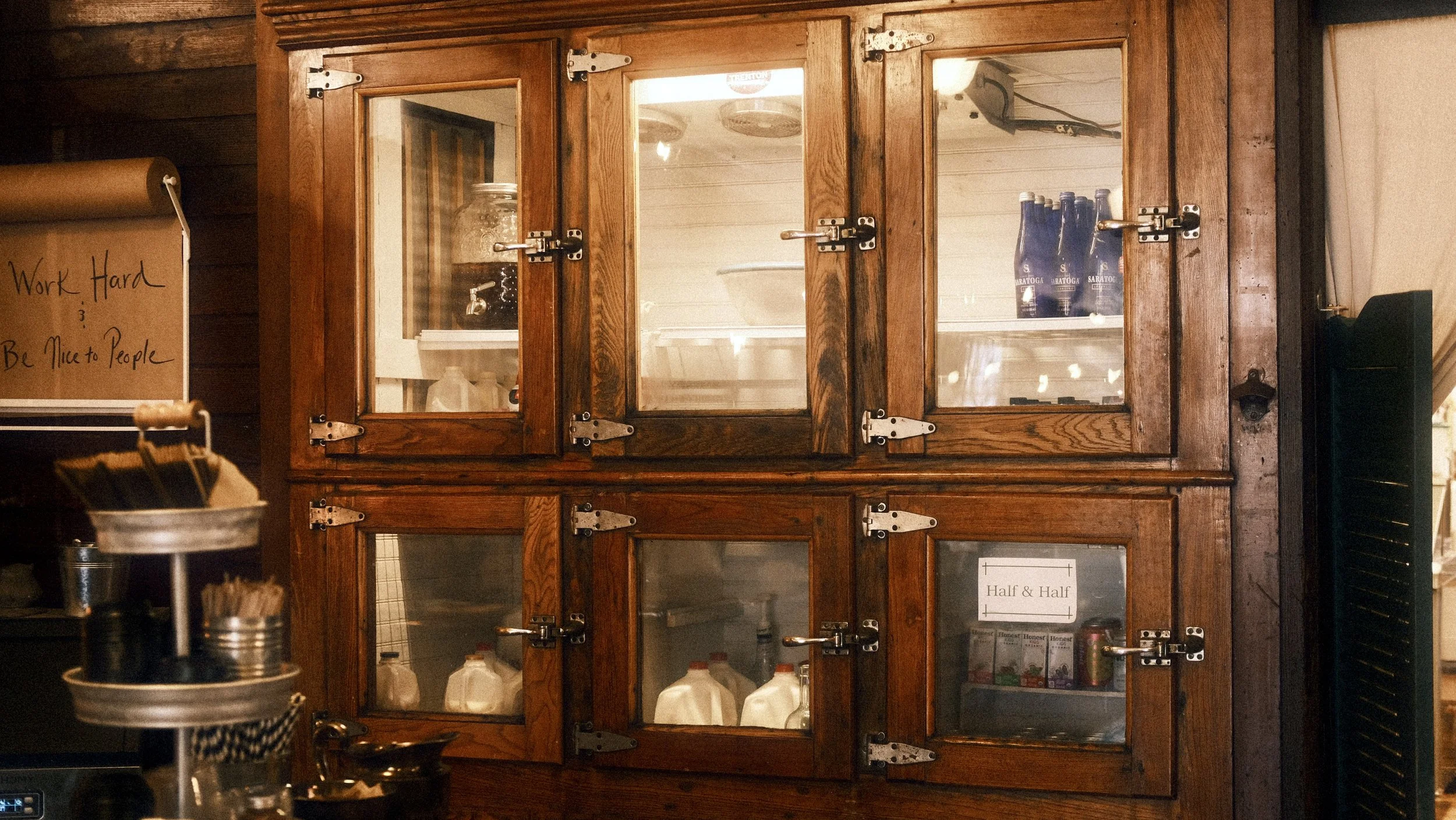 A wooden wall cabinet with glass doors, containing bottles and milk jugs inside, in a rustic setting. To the left, a brown paper-wrapped beverage dispenser and some cups are visible, with a hand-written sign on the wall that says 'Work Hard Be Nice to People'.