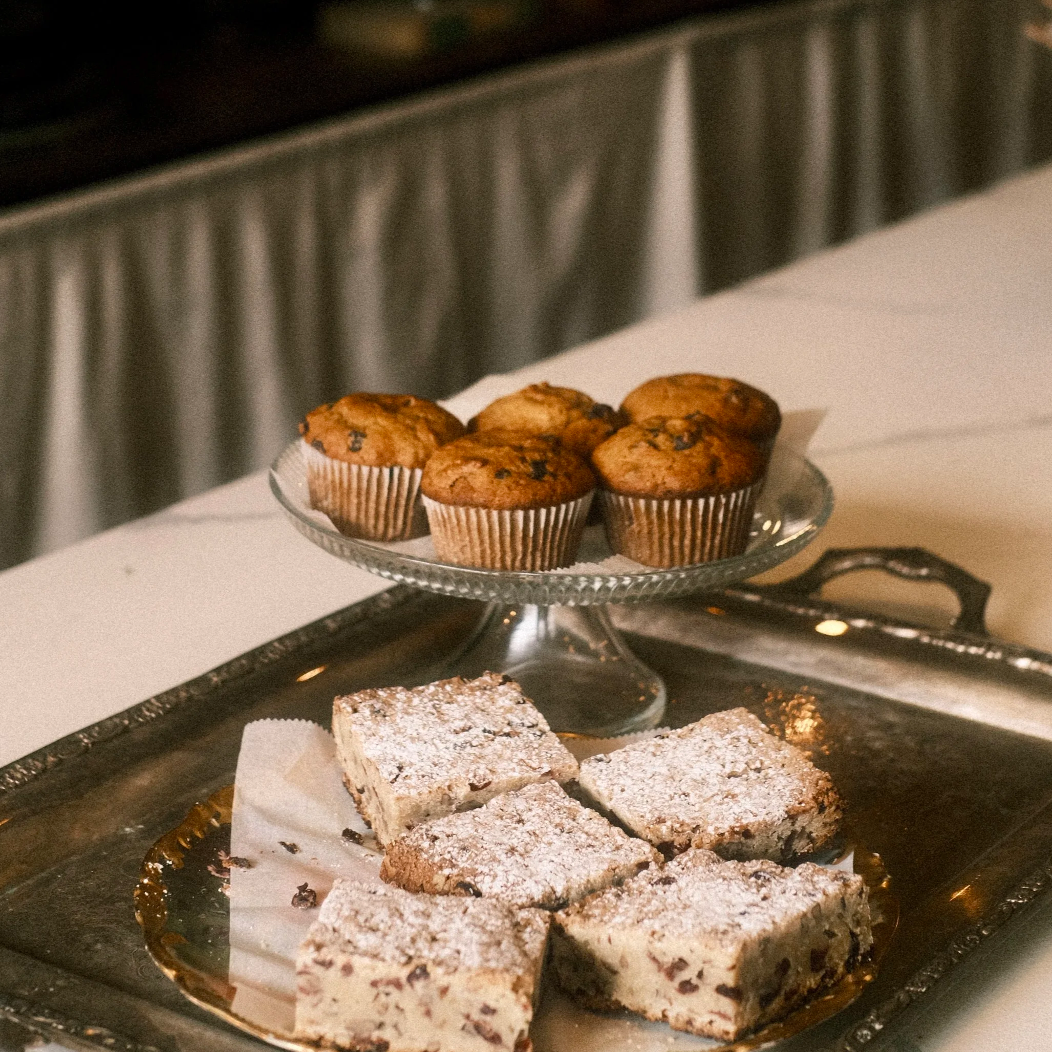 Plate of powdered sugar-covered bars and a glass dish of muffins on a white tablecloth.