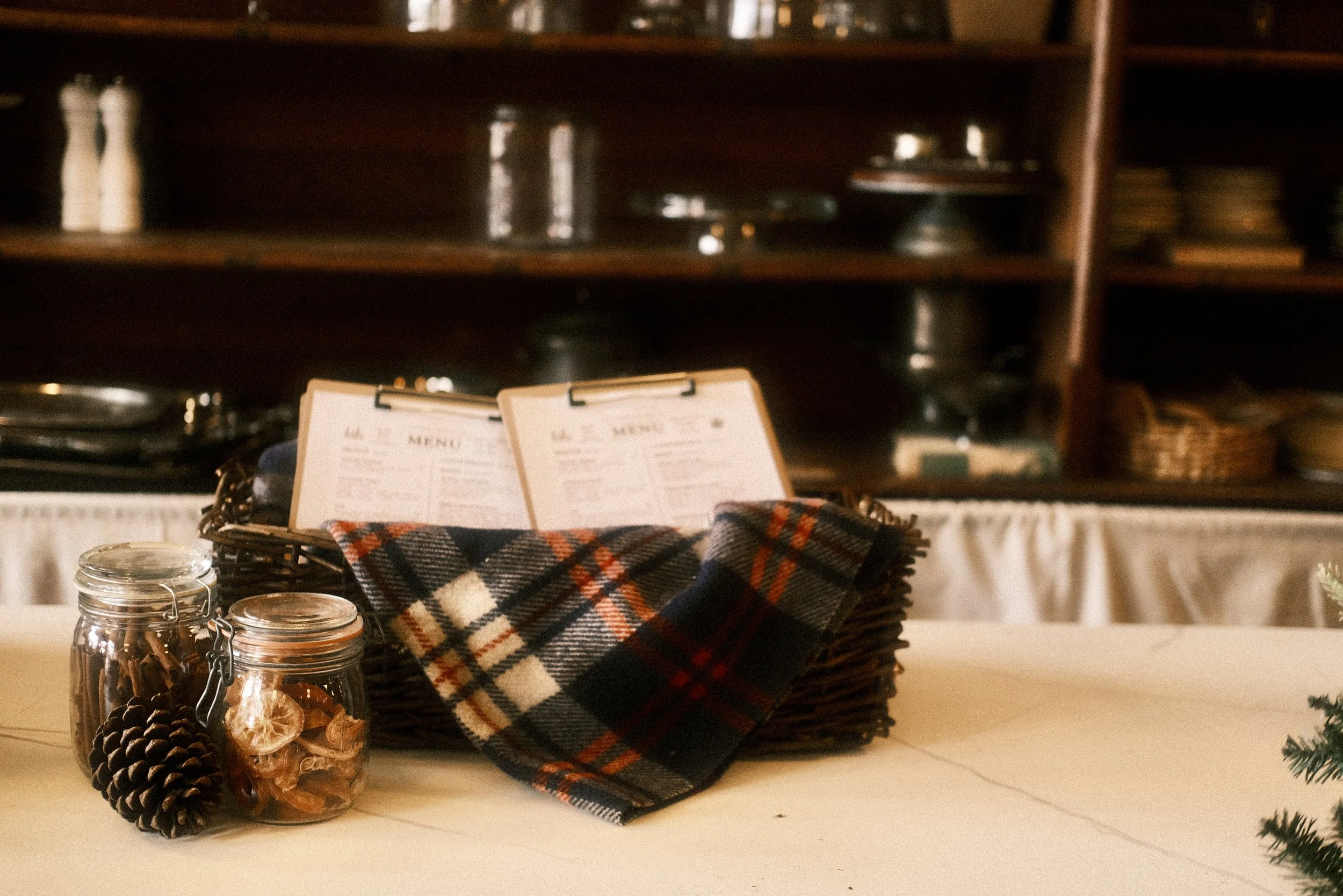 A basket with menus on top, placed on a table with two glass jars containing pinecones and dried orange slices, and a plaid cloth.