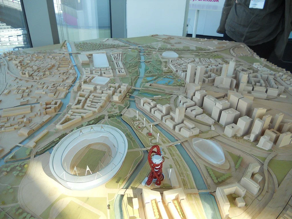 A detailed city planning model displaying various buildings, roads, waterways, and parks, with a prominent stadium and a red tower in the foreground, inside a modern building.
