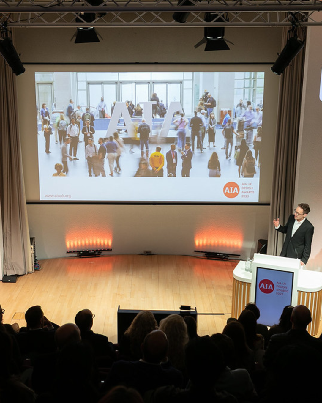 A man giving a presentation at the AIA UK Design Awards 2023, with a large screen displaying a crowded airport terminal scene with people and large letters spelling 'AIA.'